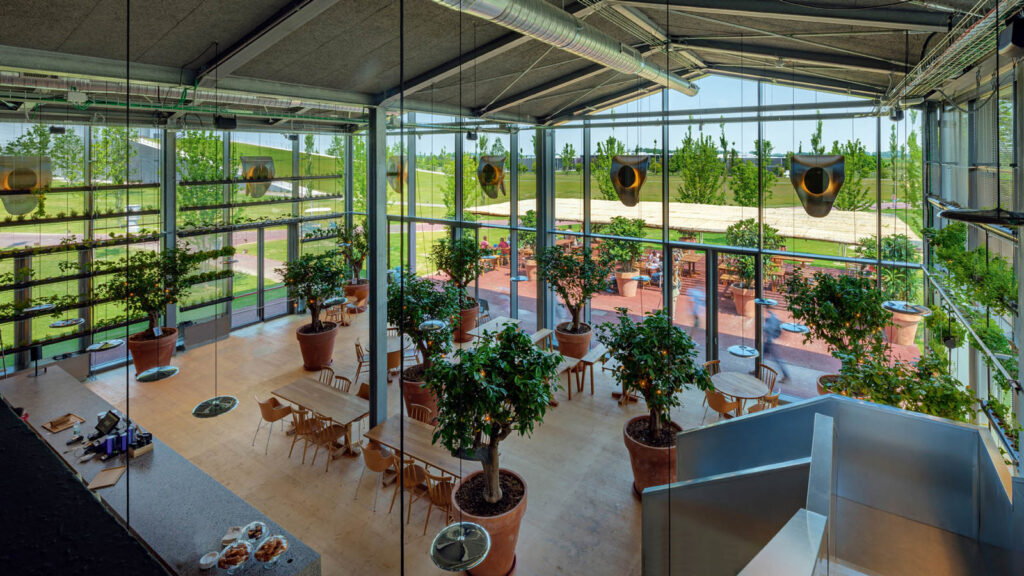 Questa vertical farm all’interno di un bar ha un interno moderno e luminoso con grandi vetrate che offrono una vista panoramica sul verde esterno dove l’erba e gli alberi ben curati creano uno sfondo pittoresco visibile attraverso le vetrate.